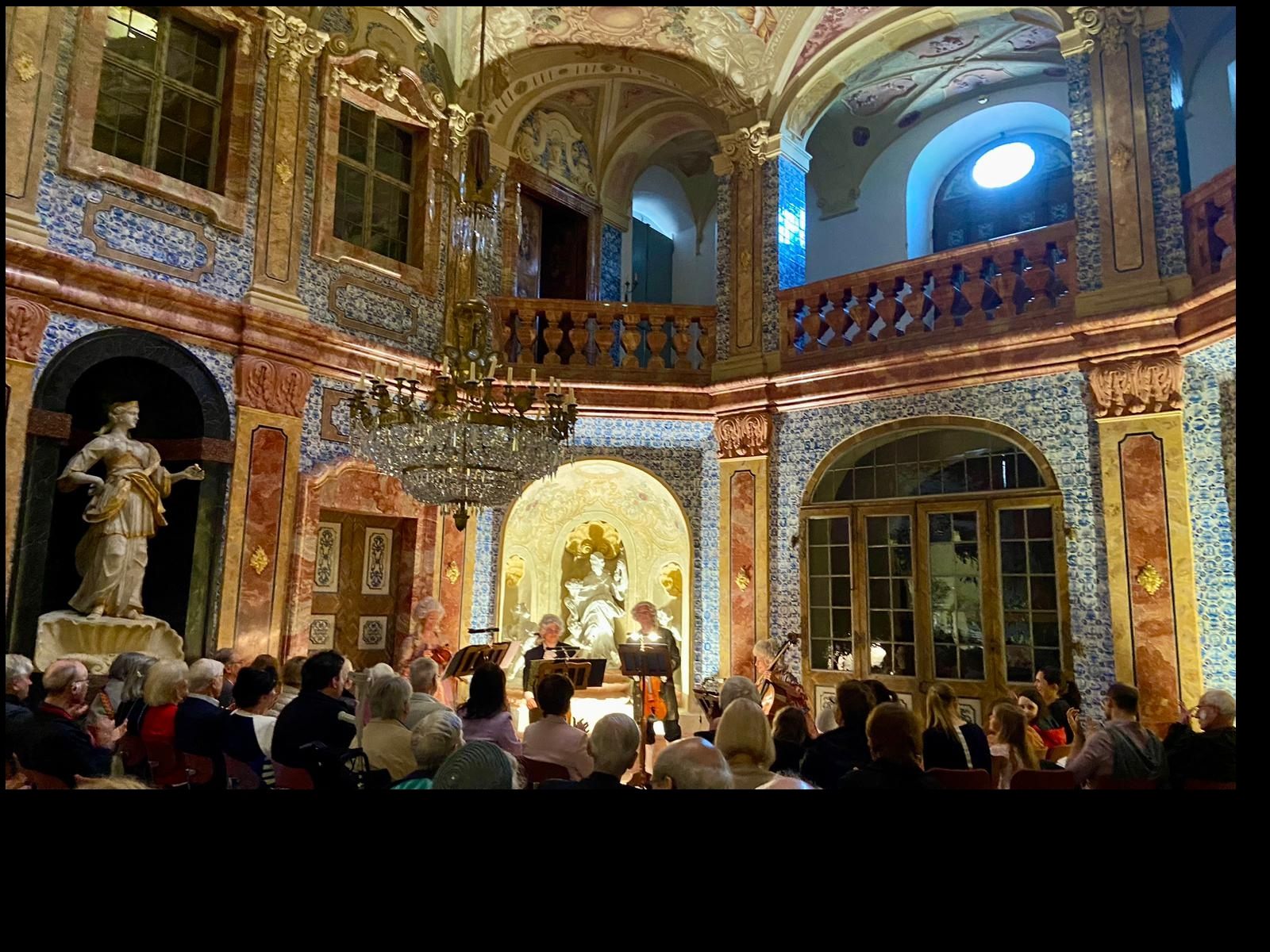 Ein Konzert in einem prunkvollen barocken Saal mit Statuen, Kronleuchter und Wandverzierungen.