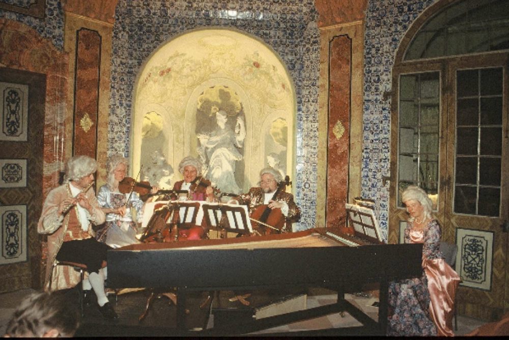 Eine barocke Musikgruppe in historischen Kostümen spielt im dekorierten Saal, mit Cembalo und Streichinstrumenten.